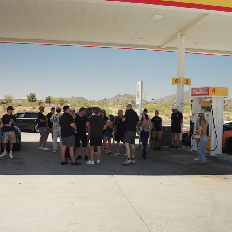 The rally group gathered under a gas station canopy at a fuel stop with the cars parked alongside.