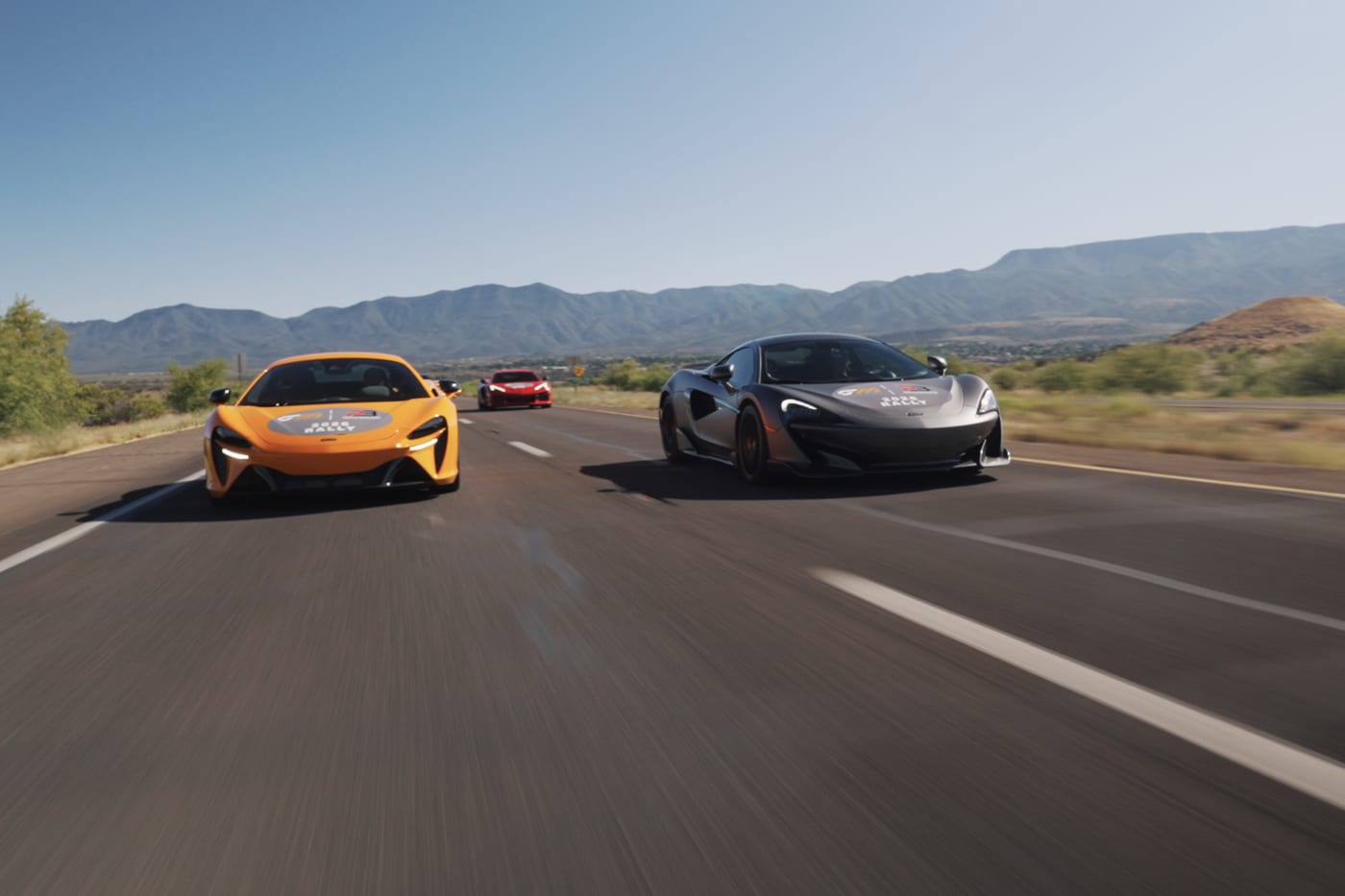 An orange McLaren GT and a grey McLaren 720S driving side by side on an open desert highway.