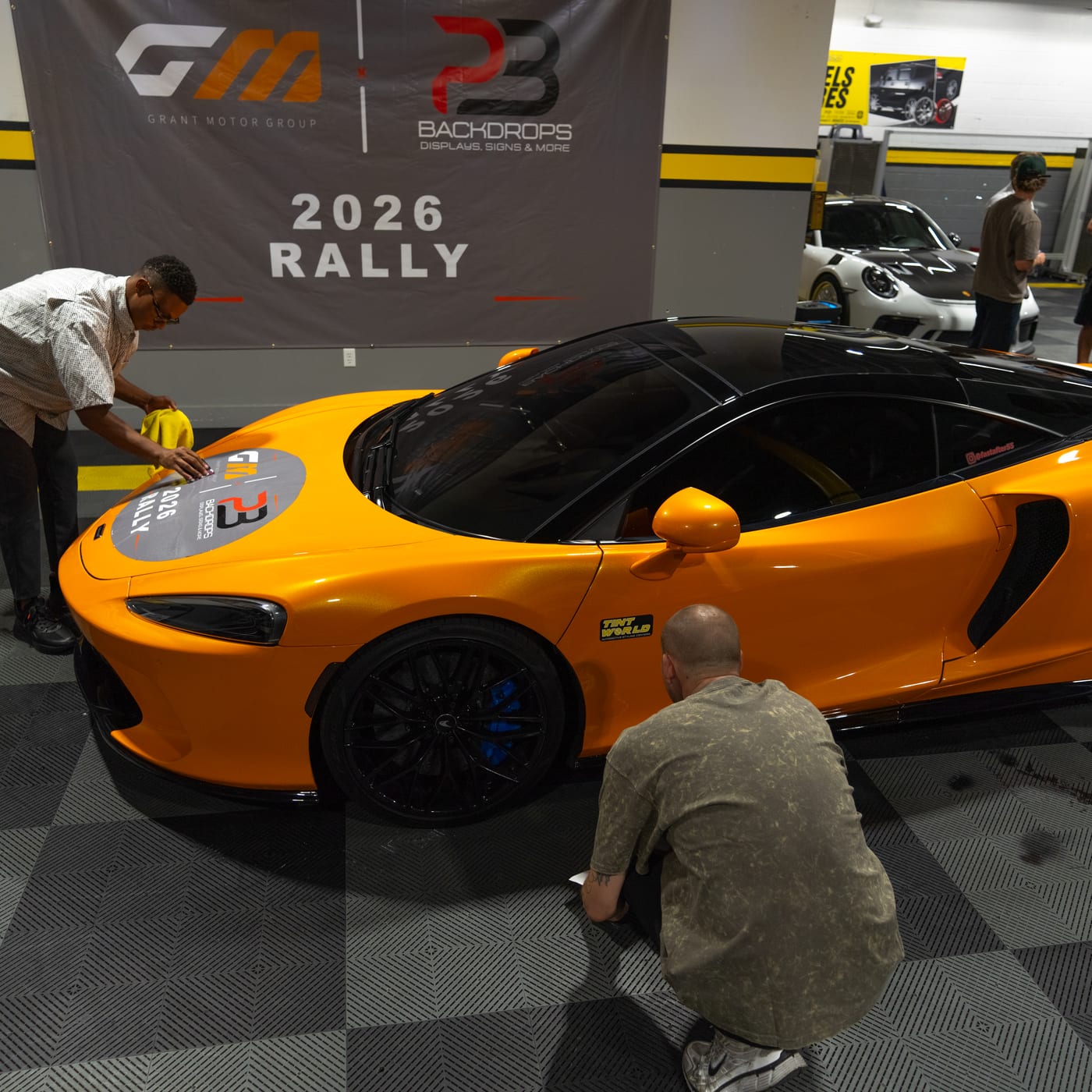 An orange McLaren GT getting a 2026 Rally hood decal applied in front of the GMG x PB Backdrops banner at the launch.
