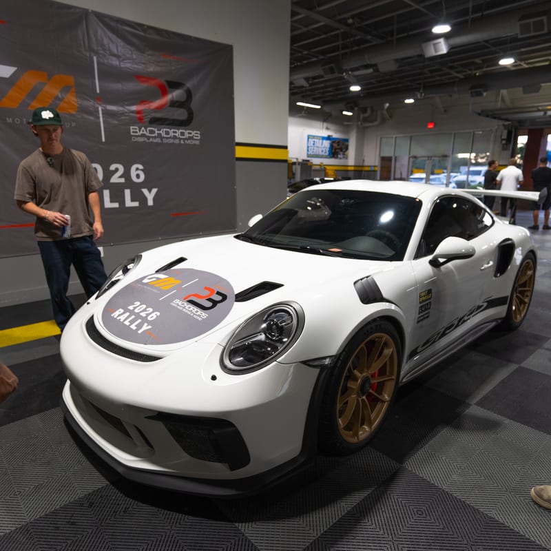 A white Porsche 911 GT3 RS with a PB Backdrops 2026 Rally hood decal parked on a checkered shop floor.